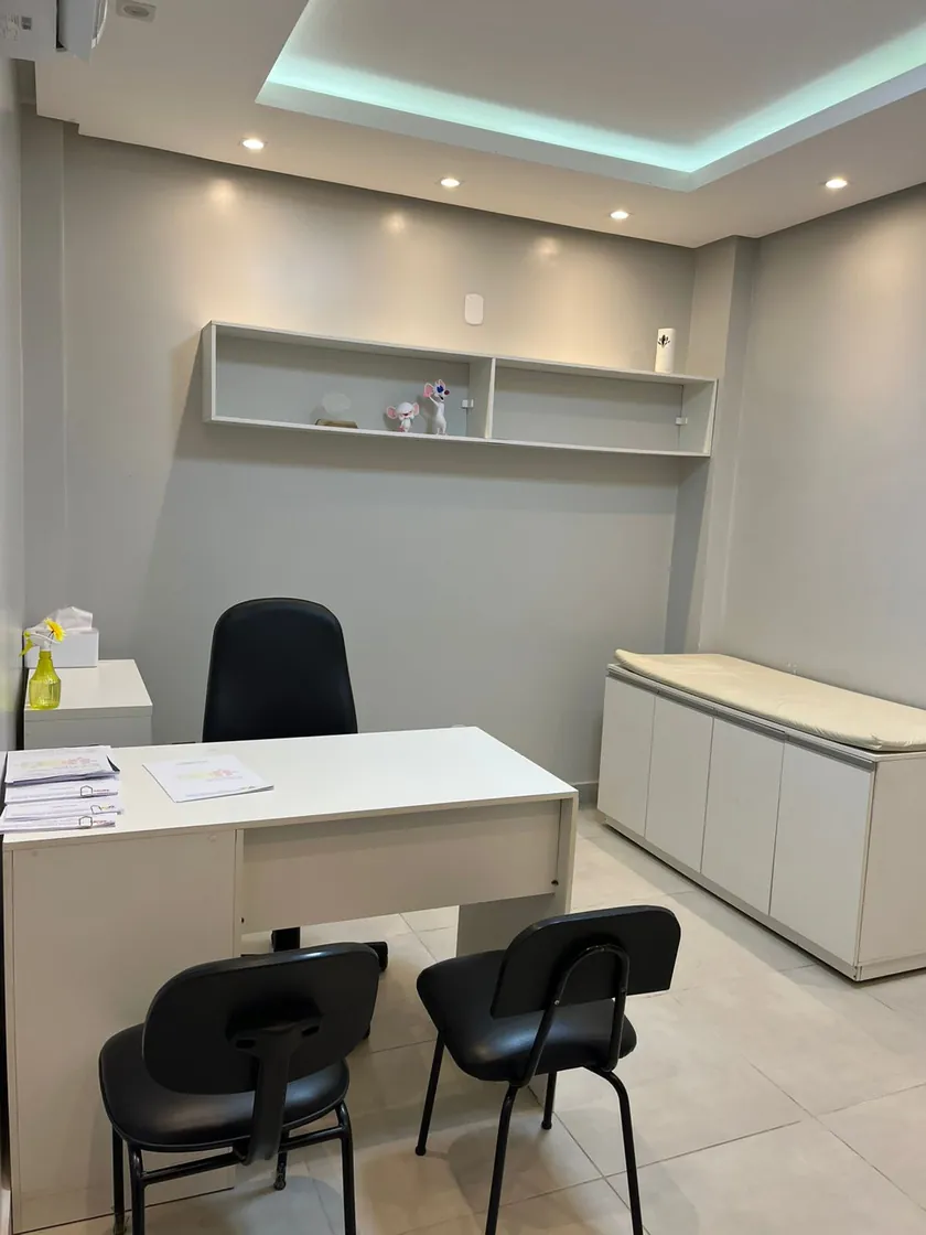 Consultório do neurologista, com mesa, cadeiras, prateleira e maca